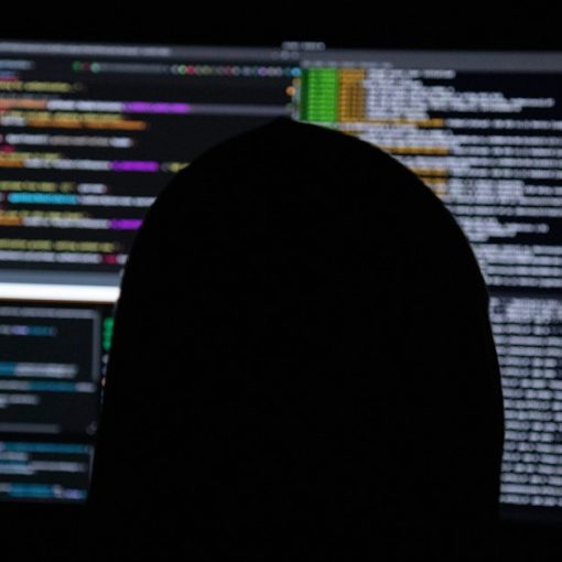 A person in silhouette against multiple computer monitors displaying lines of code, illustrating nation-state cyber wars