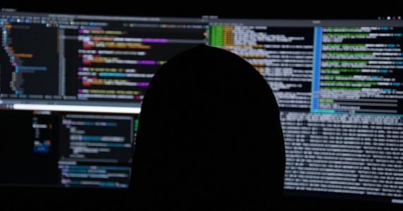 A person in silhouette against multiple computer monitors displaying lines of code, illustrating nation-state cyber wars