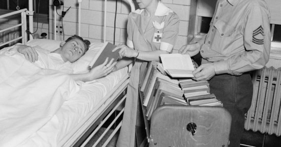 Nurse and soldier give books to patient in hospital bed, illustrating DoD desktop virtualization in healthcare