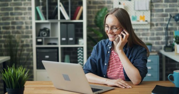 A woman talking on a cell phone while using a laptop, illustrating businesses considering VoIP