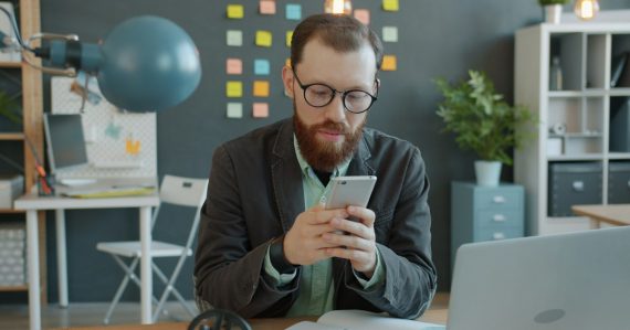 Man in glasses using smartphone at office desk, illustrating hosted PBX