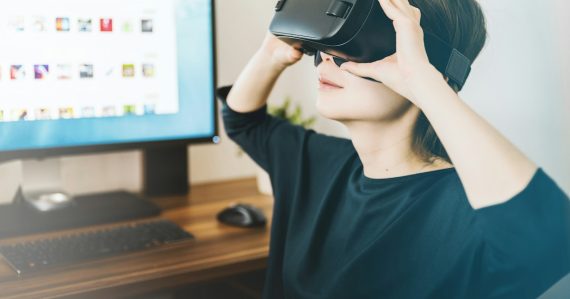 Woman using black VR headset beside computer, illustrating living with virtualization